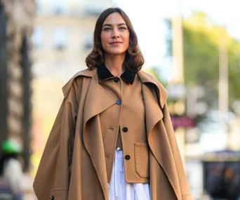 Person wearing a tan coat with black collar, standing outdoors with blurred city background.