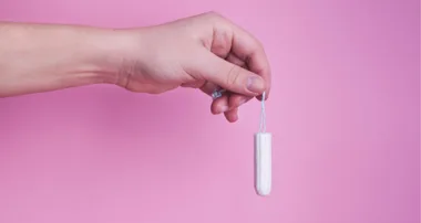 A hand holding a white tampon with a string, set against a pink background.