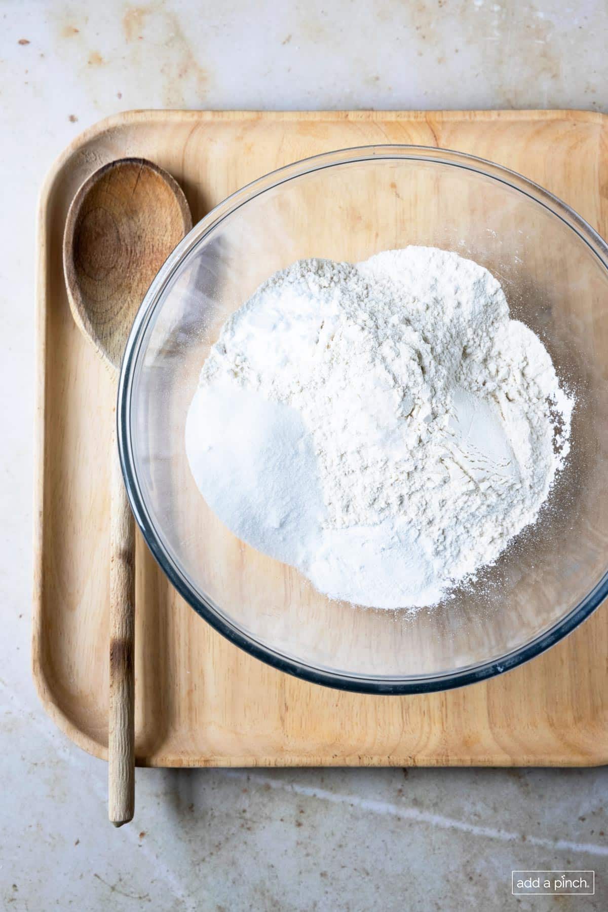 Glass bowl filled with dry ingredients used to make pancakes and a wooden spoon on a wooden tray. 