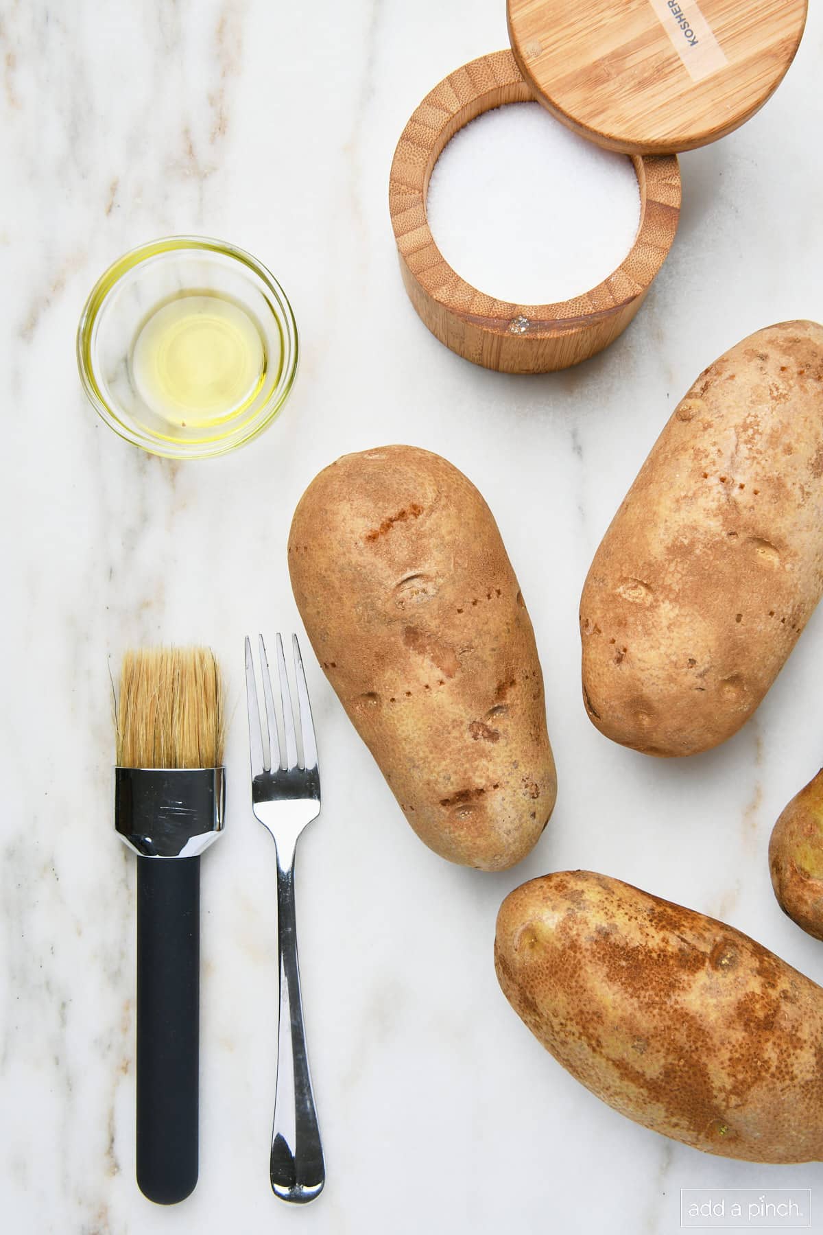 Potatoes, salt, oil, fork, and brush on a marble surface.