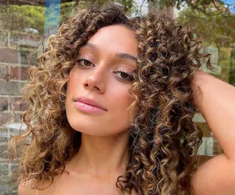 Woman with curly hair touching her head, showcasing natural beauty against a blurred brick background.