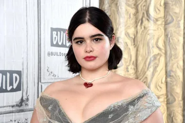 Barbie Ferreira in a lace dress with a heart necklace at a BUILD Series event, against a decorative background.