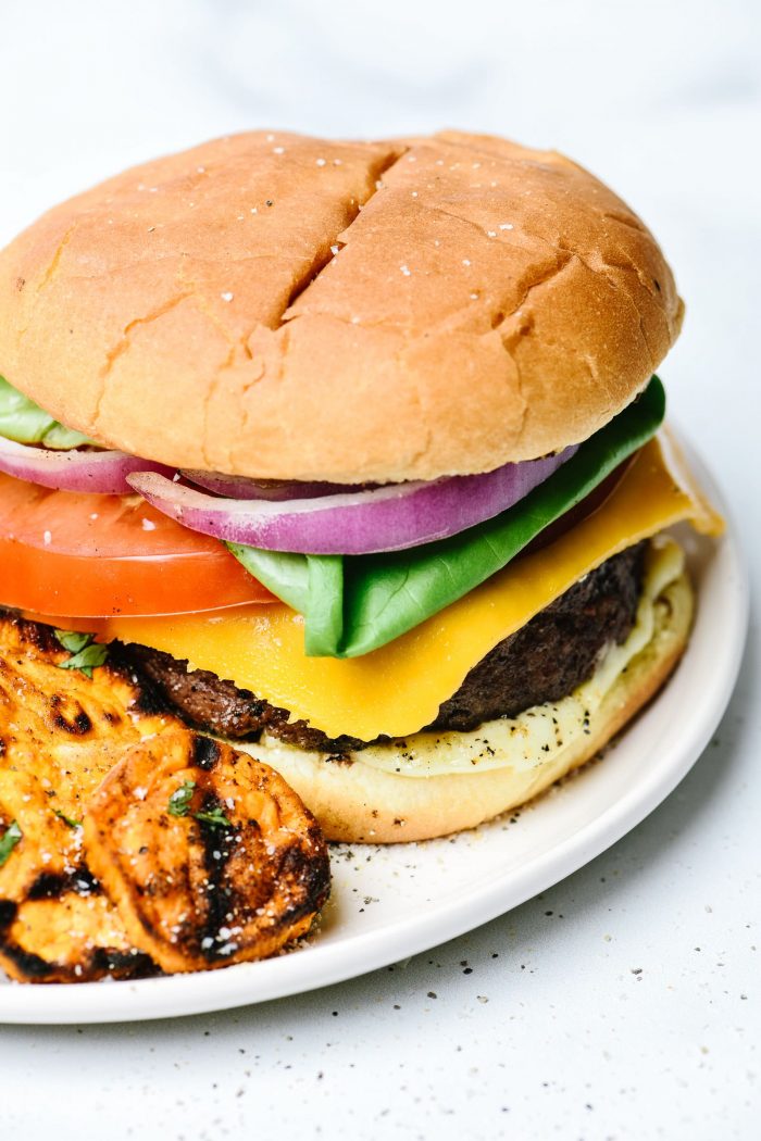 Hamburger on bun with all the toppings - onion, tomato, lettuce, cheese, mayo, served with grilled sweet// addapinch potatoes on white plate // addapinch.com