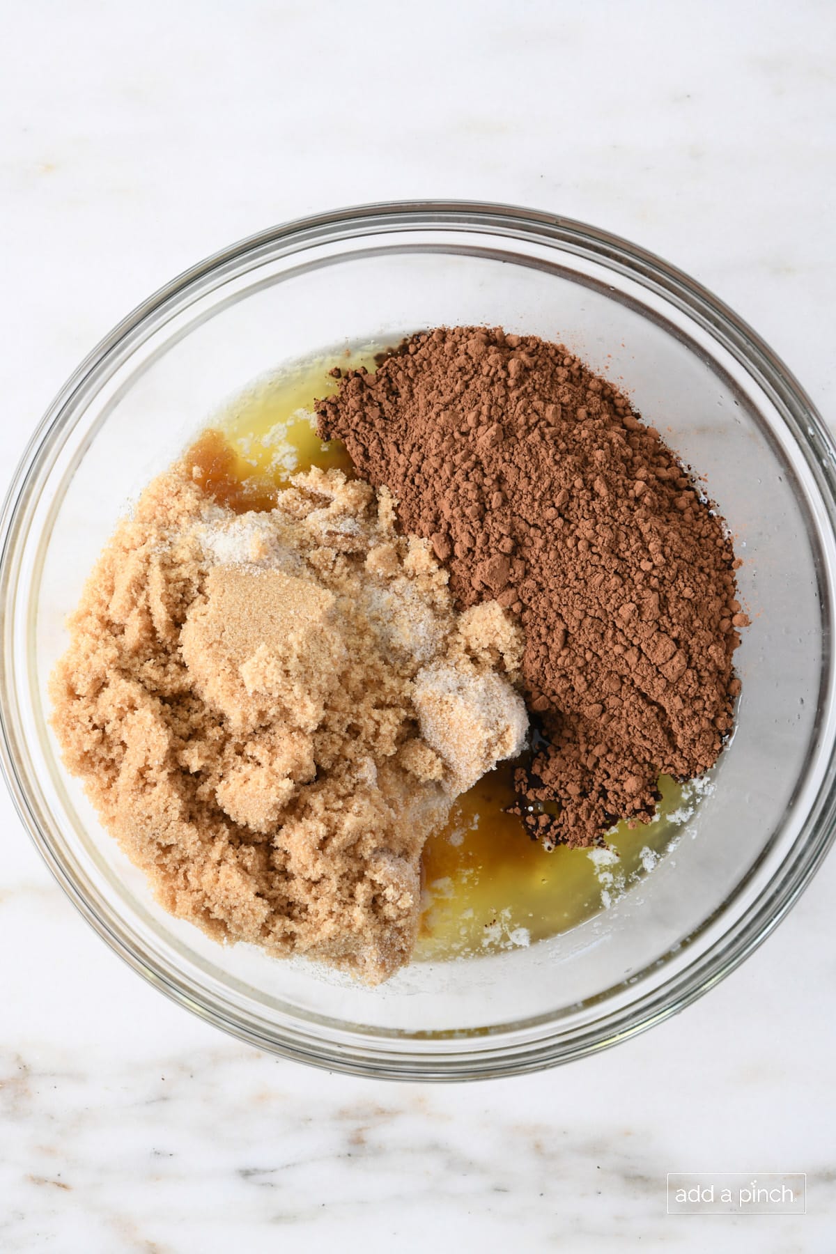 Glass bowl with melted butter, cocoa, white and brown sugar on marble counter.