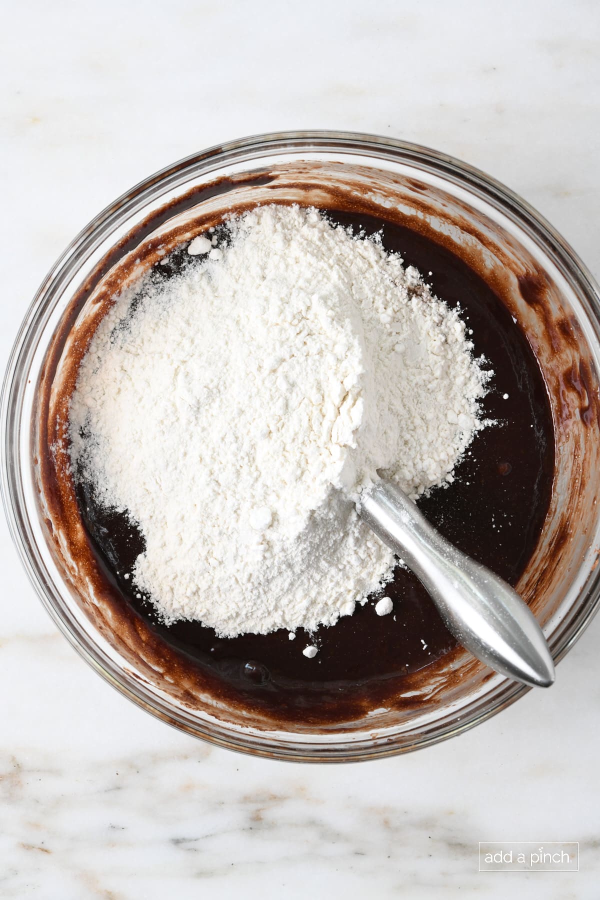 Marble countertop holds glass mixing bowl with chocolate brownie mixture with flour added ready to mix. 