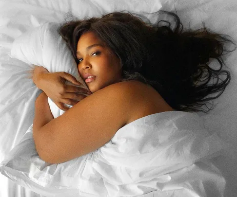 "Person lying on white bed under white sheets, looking at the camera with long dark hair spread on pillow."