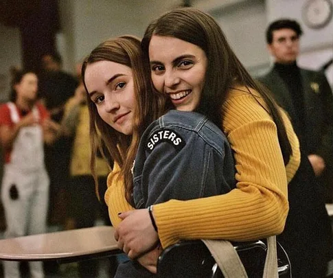 Two young women embrace in a classroom, one wearing a denim jacket with "SISTERS" text, both smiling warmly.