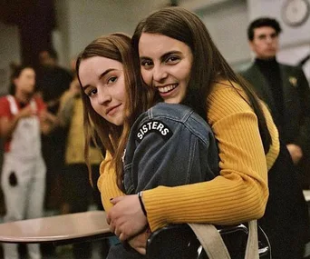 Two young women embrace in a classroom, one wearing a denim jacket with "SISTERS" text, both smiling warmly.