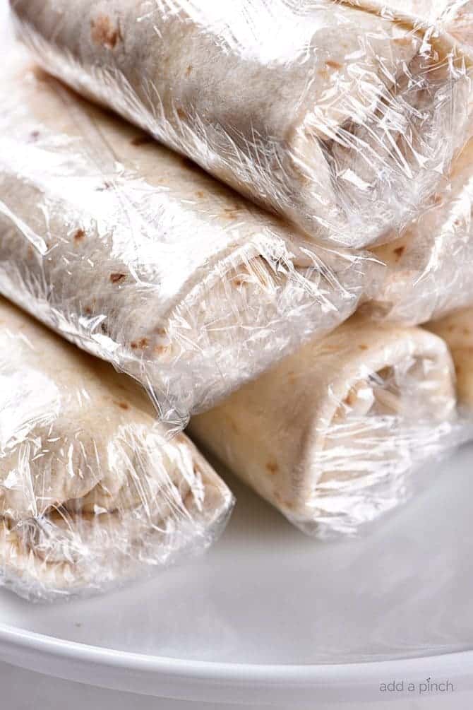 Burritos wrapped in plastic wrap on a white plate ready to be frozen.