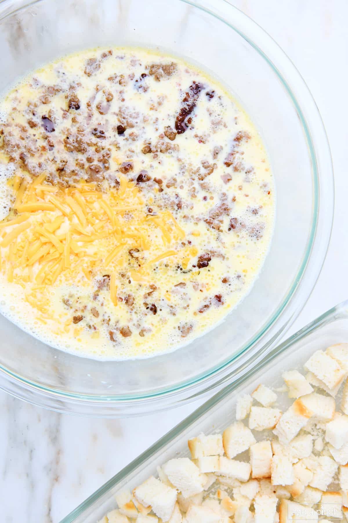 Breakfast casserole mixture in a glass mixing bowl beside casserole dish of bread cubes.