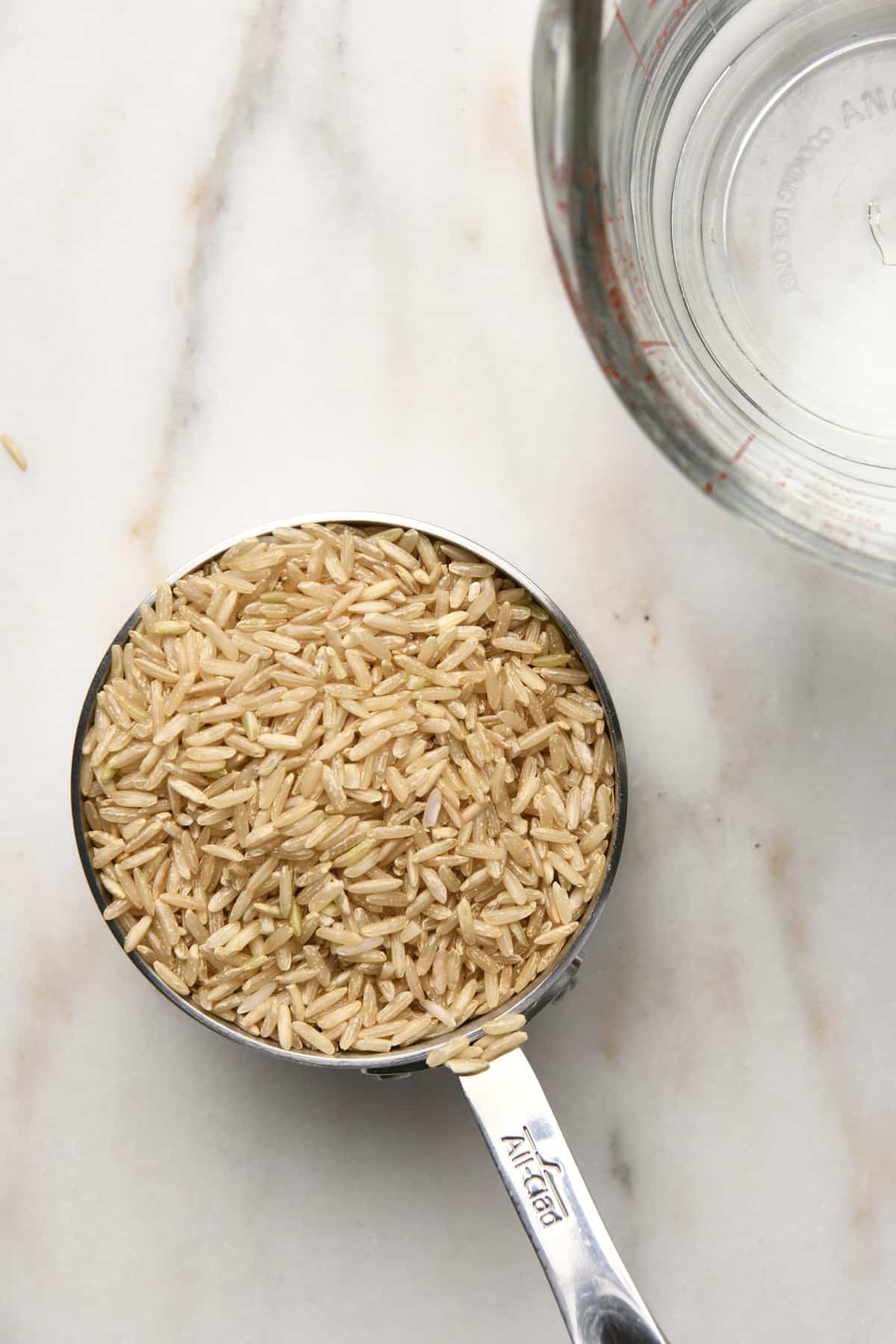 Measuring cup filled with uncooked brown rice and another with water on a marble surface.