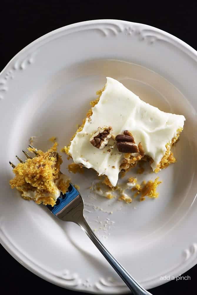 Piece of carrot cake with cream cheese frosting and toasted pecans on a white plate and dark background. | addapinch.com