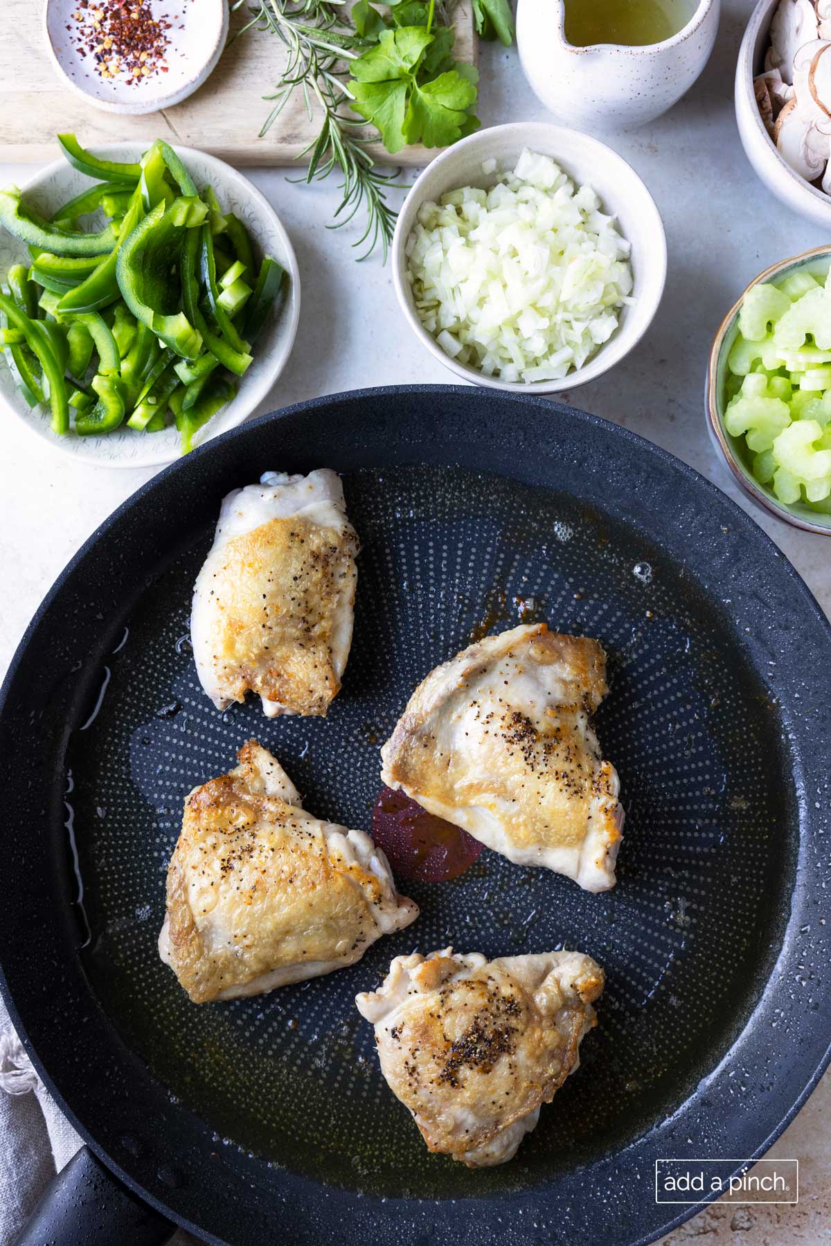 Photo of browned chicken thighs in a skillet with vegetables surrounding the skillet.