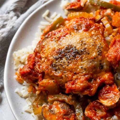 Photo of chicken cacciatore with chicken, peppers, and mushrooms on rice on a white plate.