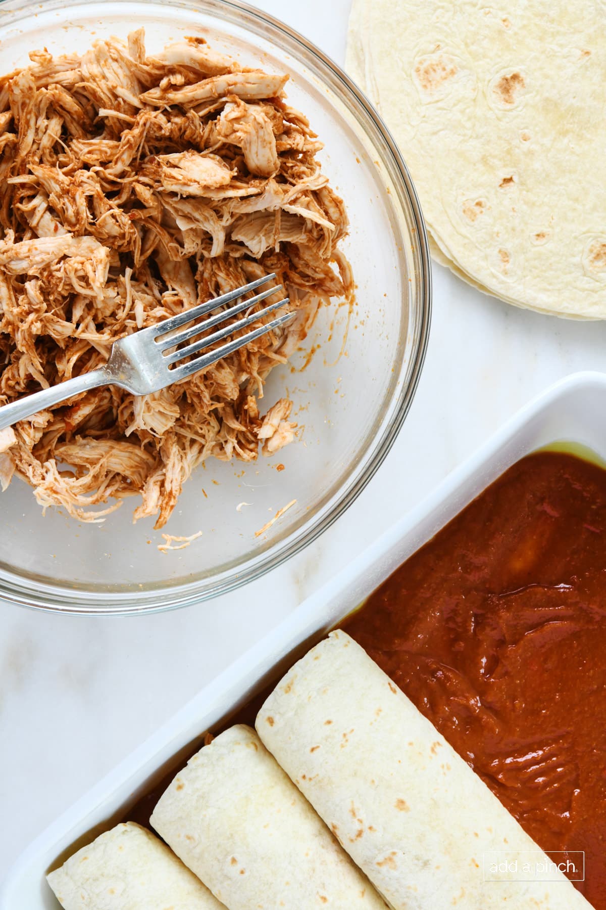 Bowl of shredded chicken, stack of tortillas, and a baking dish with some assembled enchiladas.