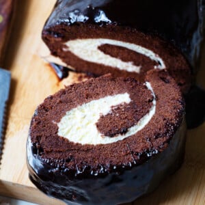 Slice of chocolate cake roll with the rest of the cake showcasing the cream filling and the roll design in the cake.