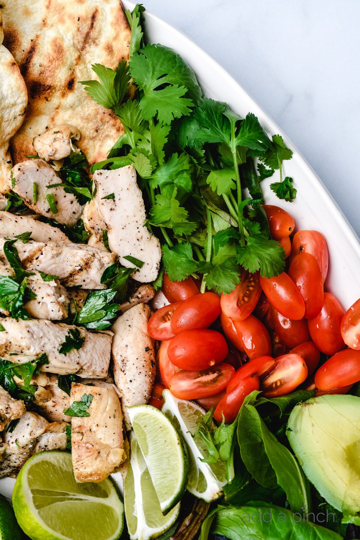 Grilled chicken, tomatoes, limes, avocado, fresh cilantro, and grilled bread on a white platter.