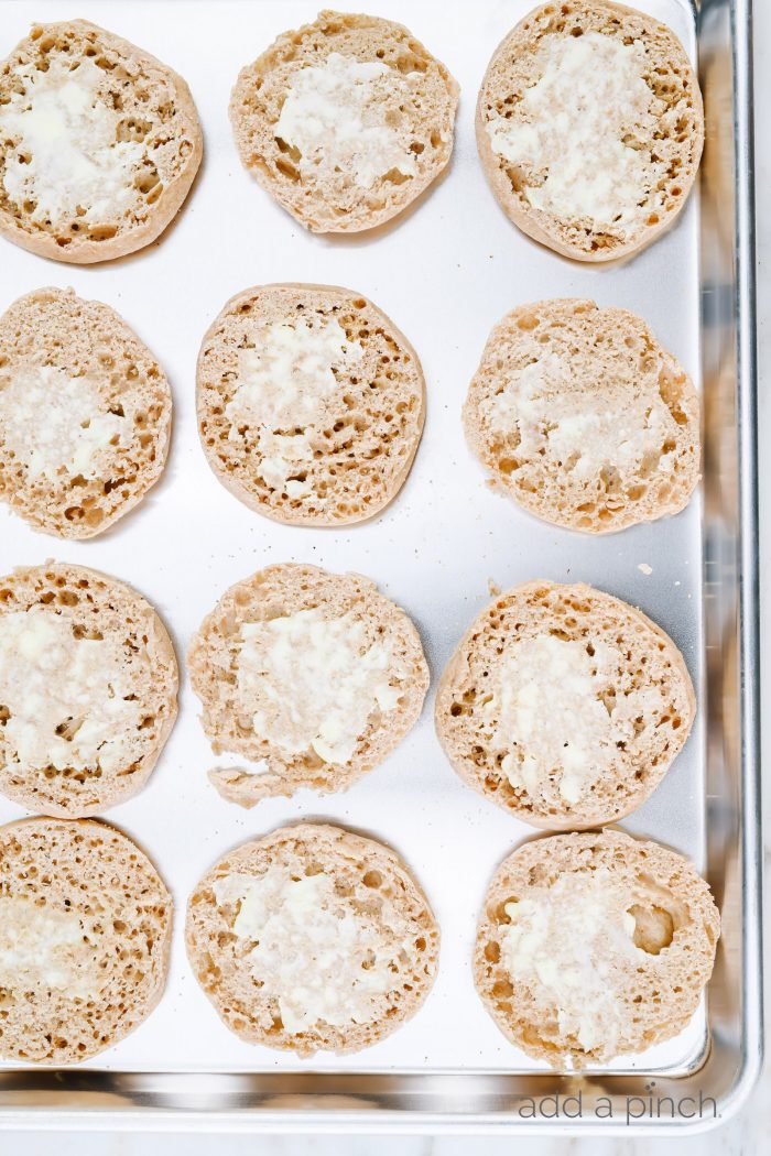 Baking sheet with buttered halves of English muffins.