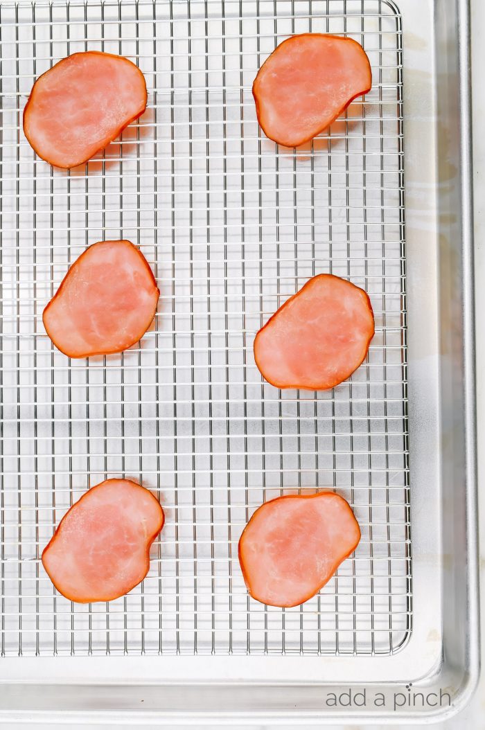 Six slices of Canadian bacon are on a wire rack fitted baking sheet.