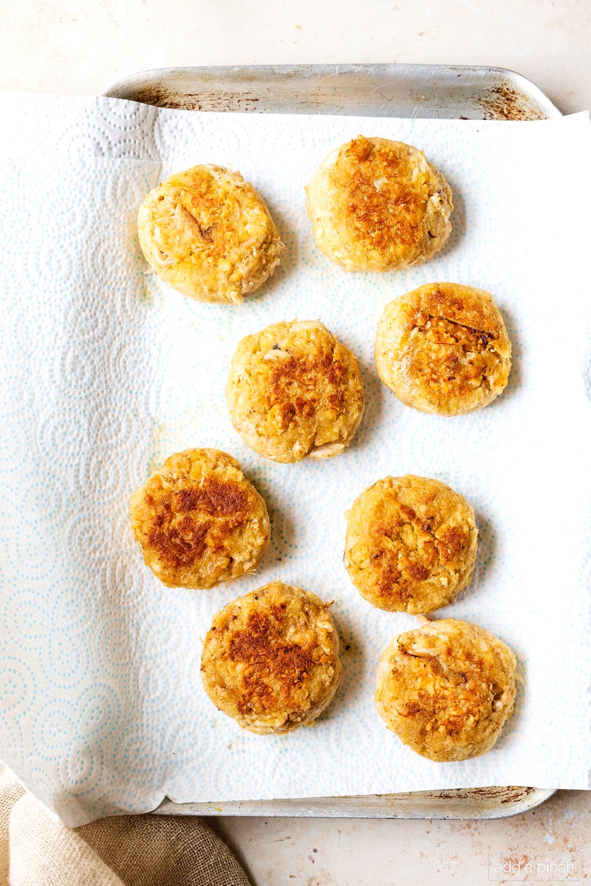 Baking sheet lined with paper towels has 8 cooked crab cakes to drain.