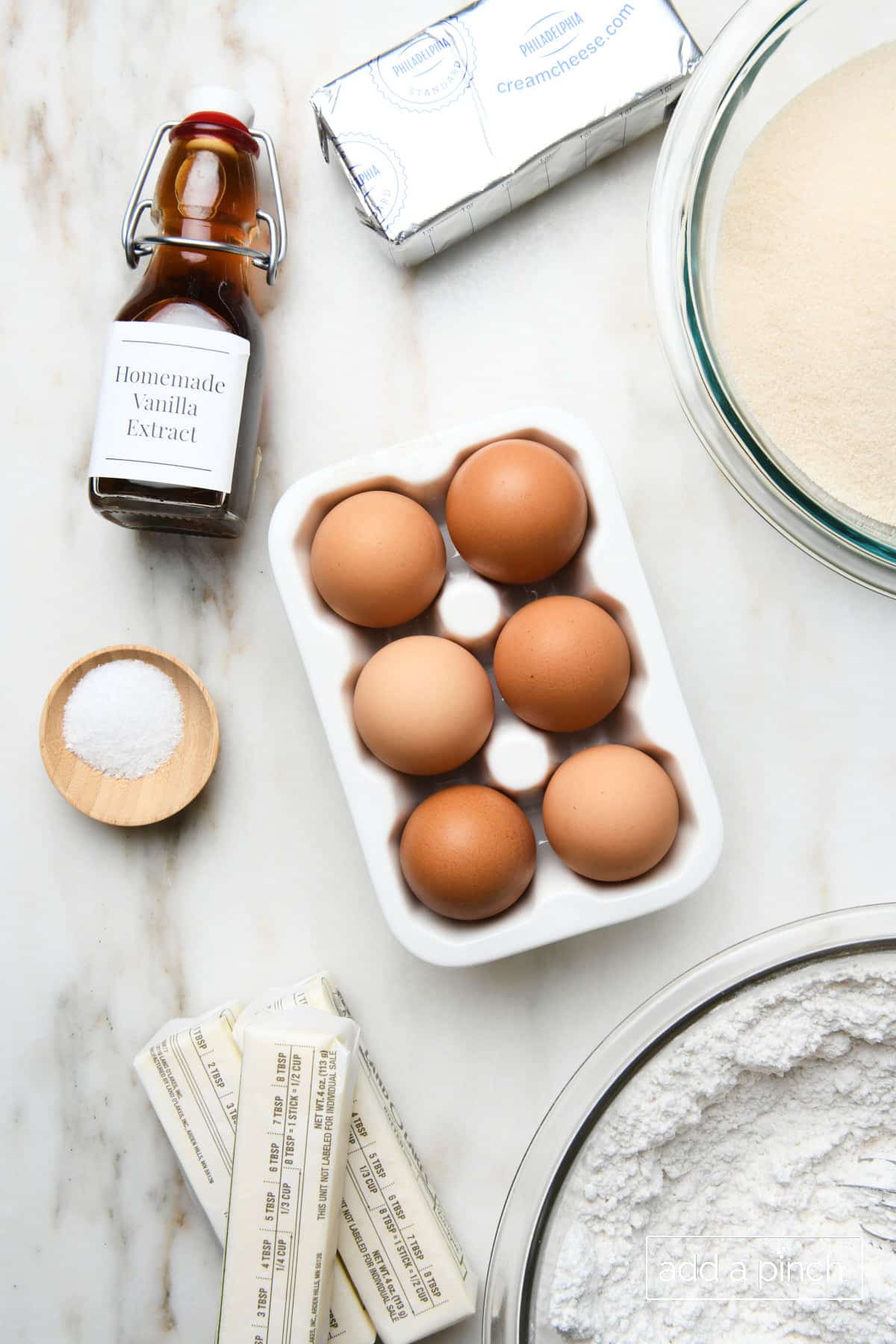 Sugar, block of cream cheese, six eggs, homemade vanilla extract, flour,  three sticks of butter, and salt all on a marble counter. 