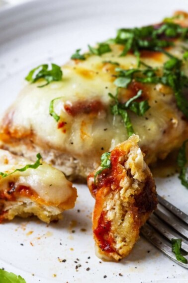 Photograph of chicken parmesan recipe with a bite on a fork served on a white plate.