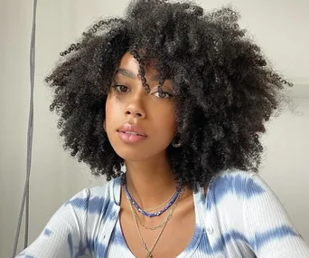 Young woman with voluminous curly hair wearing layered necklaces and a tie-dye shirt, posing indoors.