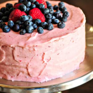 Strawberry cake with strawberry buttercream frosting topped with fresh blueberries and strawberries on a silver cake stand.