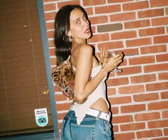 Woman in jeans and tank top holding a wine glass, standing against a brick wall with a leopard print bag.