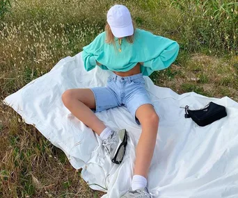 Person in mint green crop top, denim shorts, and cap sitting on a blanket outdoors.