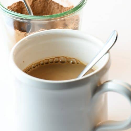 Photograph of white mug filled with hot cocoa and a clear jar of hot cocoa mix behind it on a white counter. // addapinch.com