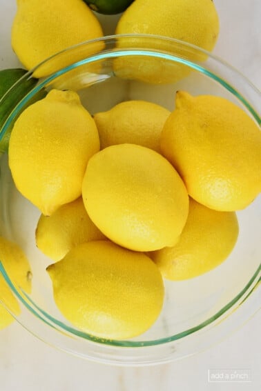 photo of glass mixing bowl filled with yellow lemons and limes on the side of the bowl on a marble surface.