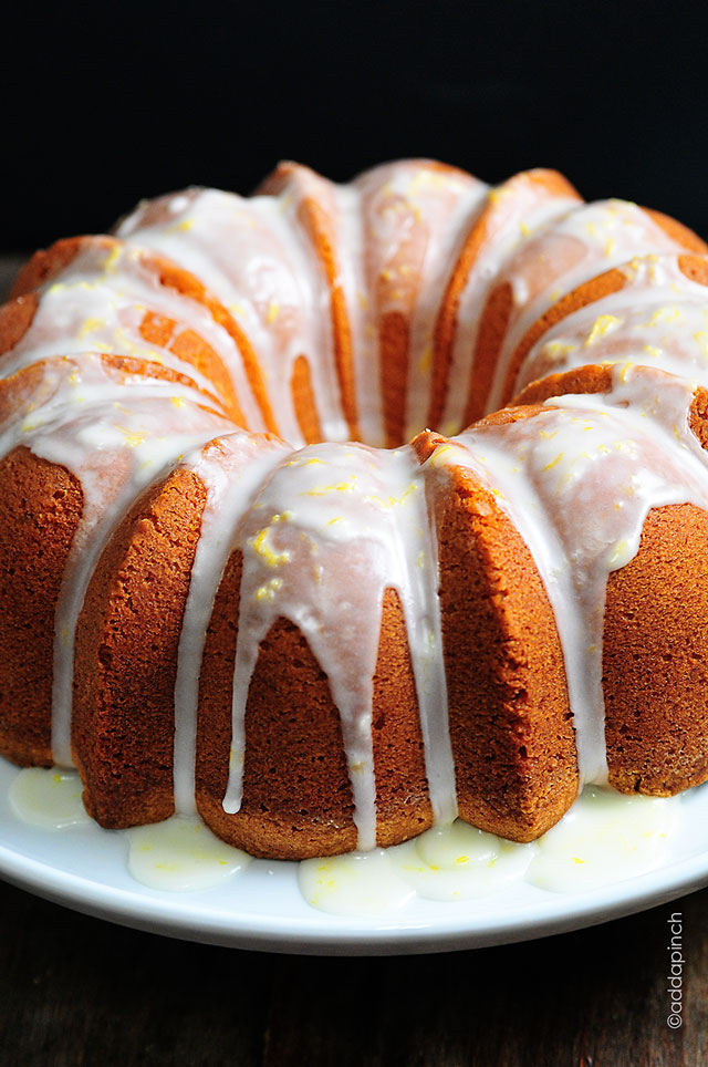 White plate hold Lemon pound cake drizzled with glaze that shows lemon zest in it. ©addapinch.com