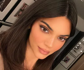 Brunette woman with natural makeup, full lashes, and soft lip color, posing in a room with framed photos on the wall.