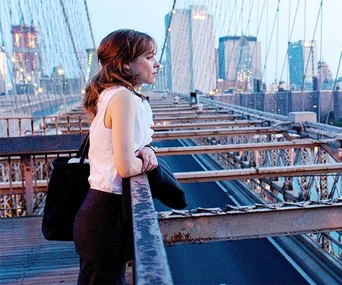 A woman leans on a bridge railing, gazing at the city skyline in the evening light.