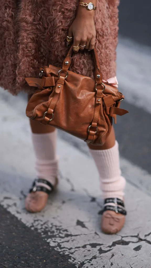 A Fashion Week guest is seen wearing a long fluffy rose vest, several chunky rings in gold and silver, a gold watch, a rectangular brown leather bag with silver details, pale pink leg warmers, and beige leather ballet flats with small bows and two black leather straps featuring silver buckles and eyelets from Miu Miu before the Miu Miu Womenswear Spring/Summer 2025 Show as part of Paris Fashion Week on October 01, 2024 in Paris, France.