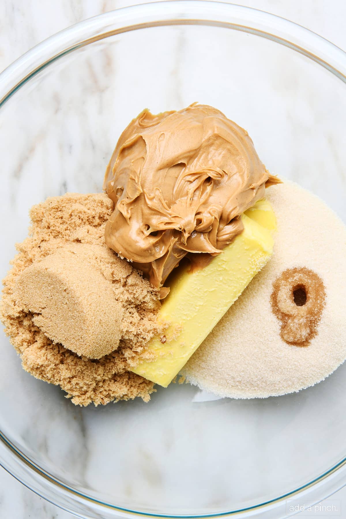 Peanut butter, brown sugar, sugar, butter, and vanilla extract in a mixing bowl on marble surface.