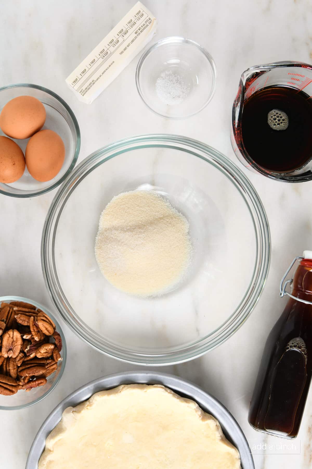 Mixing bowl with sugar, unbaked pie crust, vanilla extract, pecan halves, three eggs, salt, butter, and measuring cup with maple syrup. 