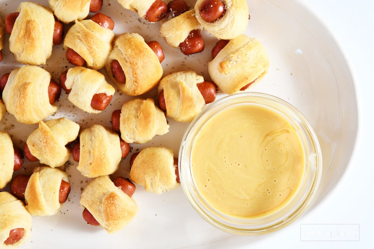 White platter filled with cocktail sausage wrapped crescent rolls and a honey mustard dipping sauce.