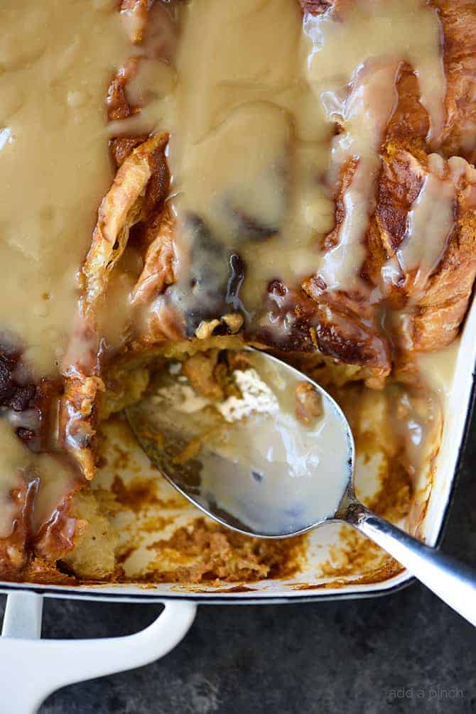 Spoon in white baking dish has scooped out serving of pumpkin bread pudding with maple cream sauce. 