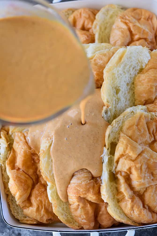 White baking dish with sliced croissants and pumpkin pudding being poured onto it.