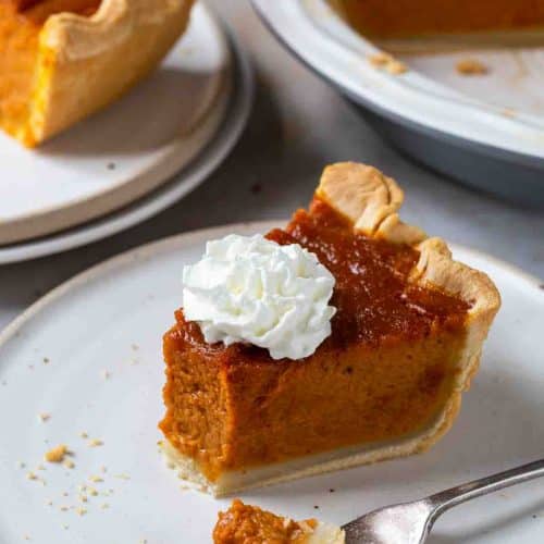 Slice of pumpkin pie with one bite on a fork. Remaining pie in the background. All on white surfaces.