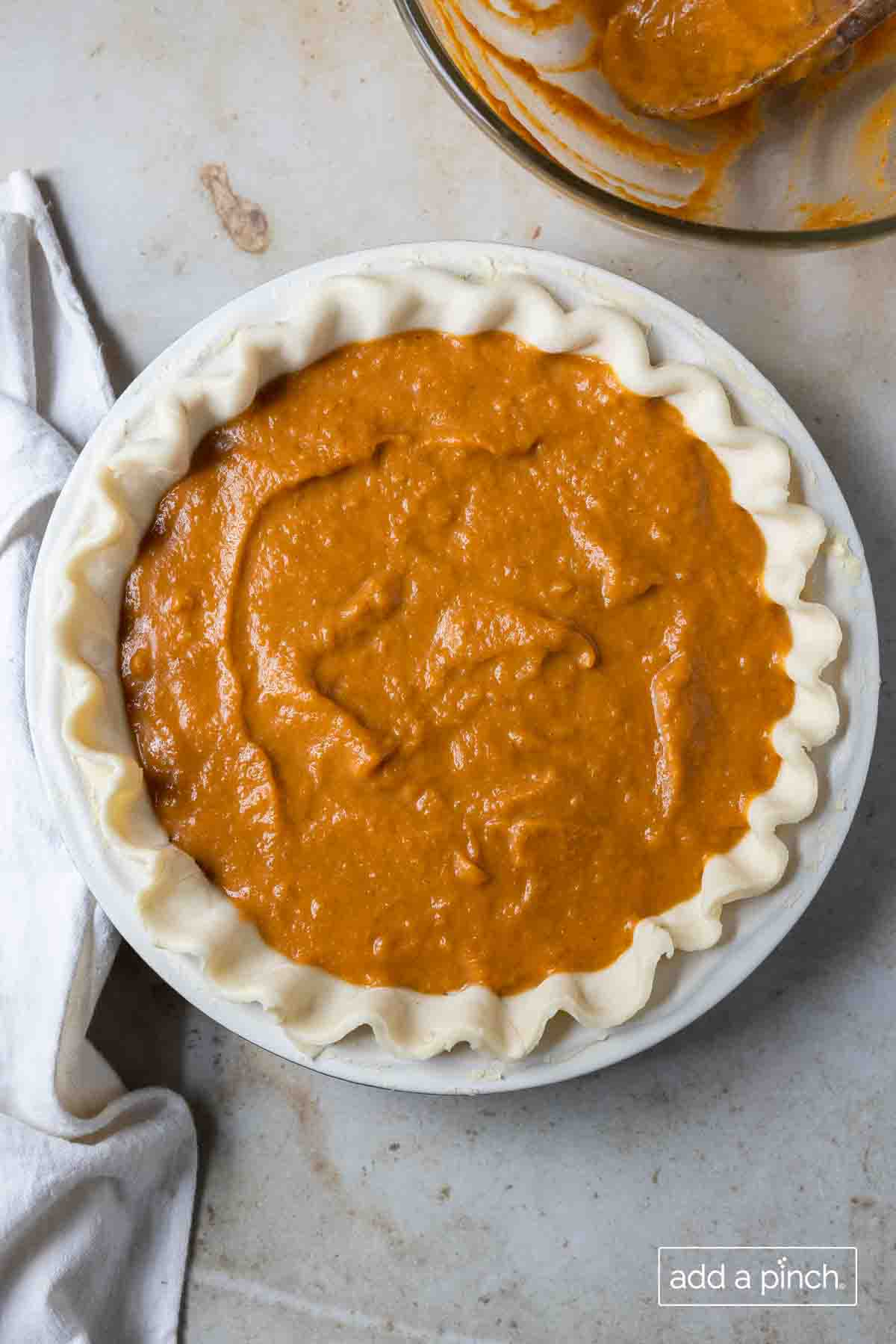 Unbaked, fluted pie crust filled with pumpkin pie filling in a white pie plate on a light stone counter.