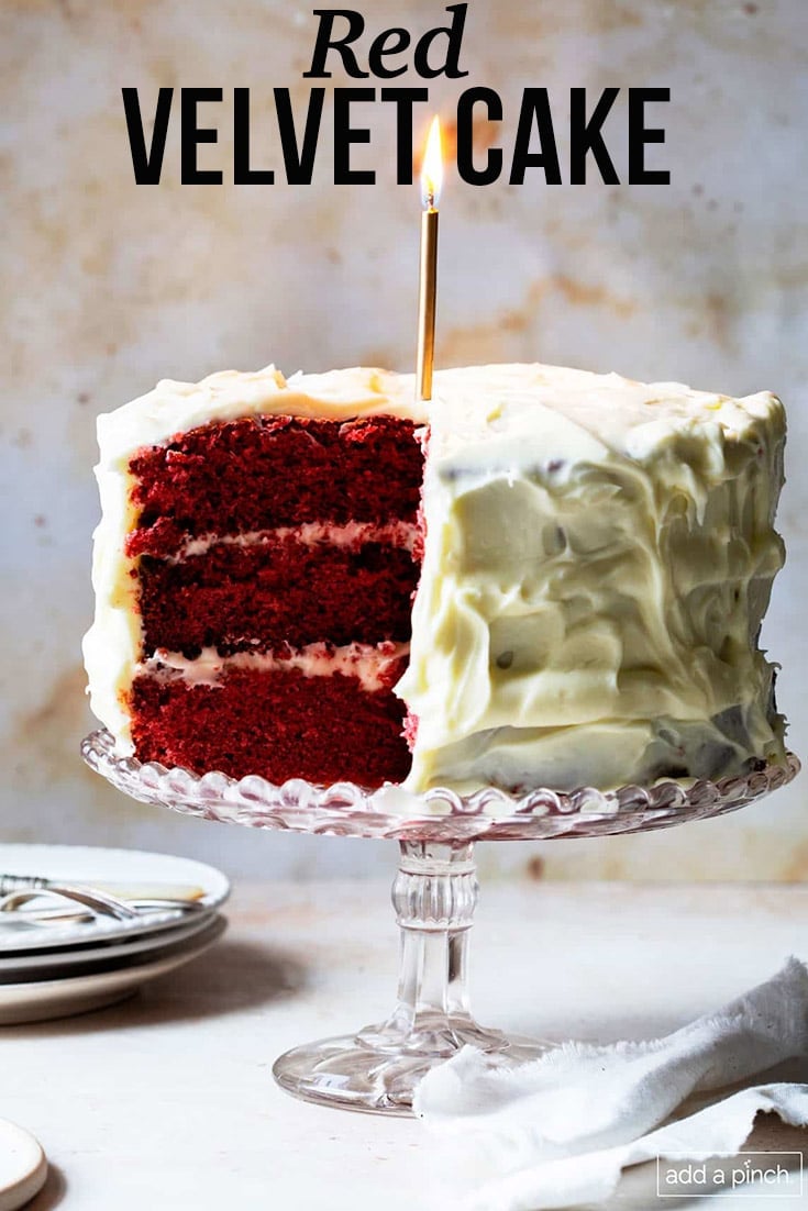 Red Velvet Cake with Heritage Frosting topped with a single gold candle on a crystal cake stand and stack of plate to side - with text - addapinch.com