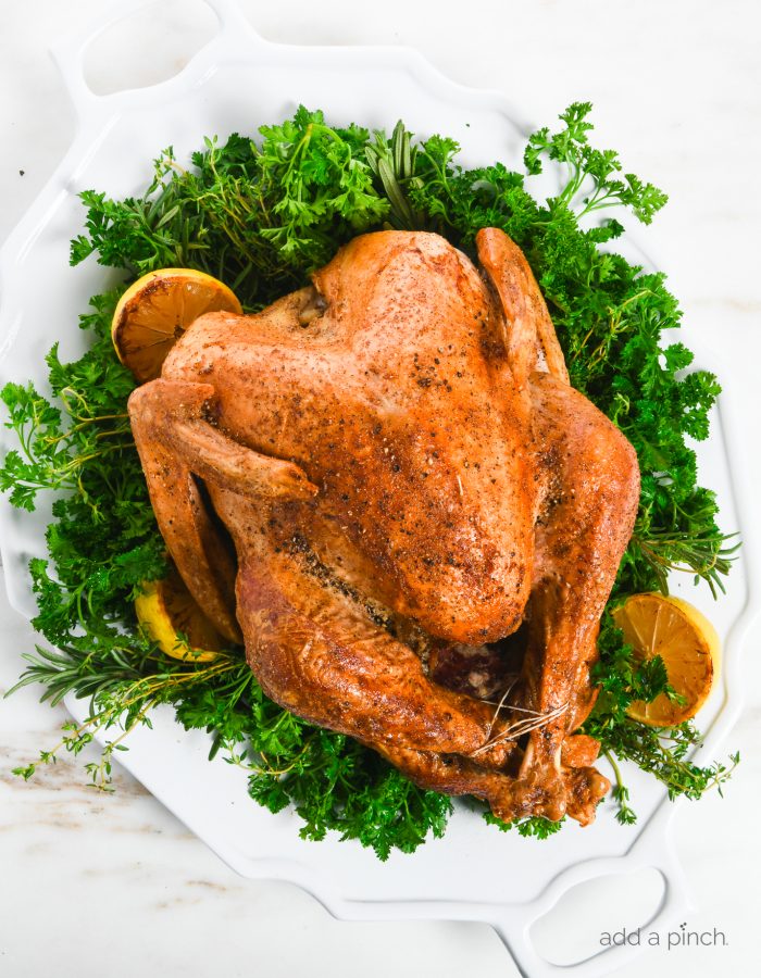 Overhead photo of a golden brown turkey on a large white platter garnished with fresh herbs, lemon, salt and pepper.