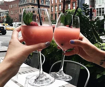 Two hands toasting with glasses of pink rosé slushies garnished with mint leaves and strawberries, in an urban outdoor setting.