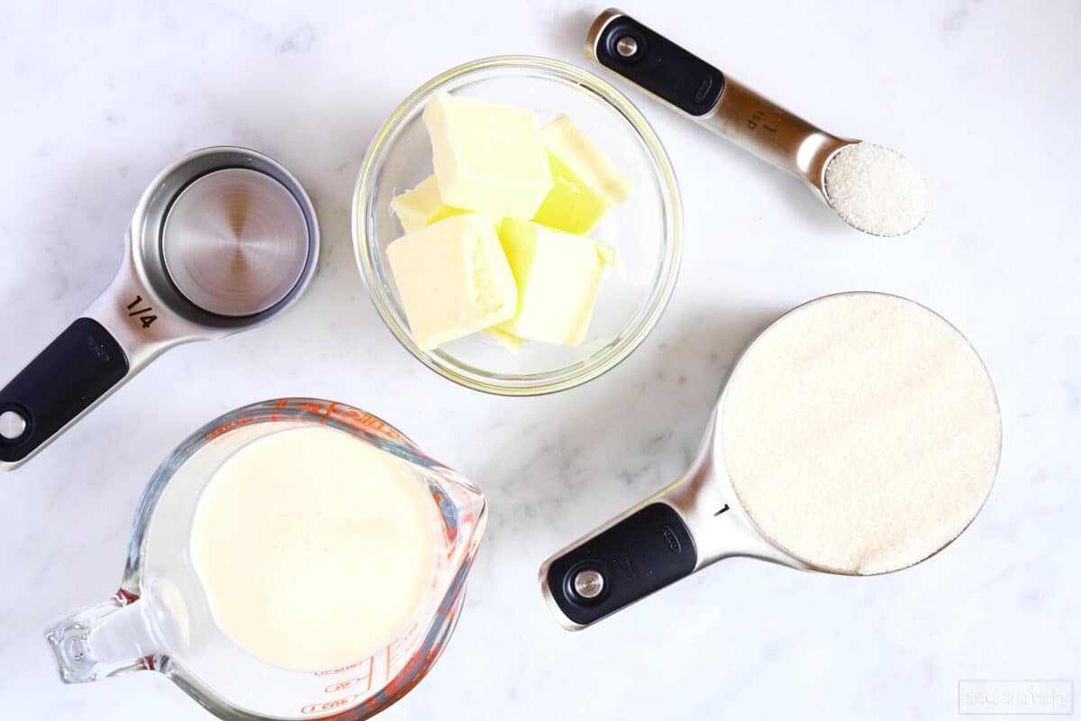 Ingredients used to make caramel sauce (sugar, butter, heavy cream, water, and salt) on a marble counter.