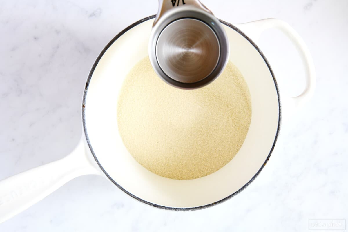 Water in a measuring cup being added to sugar in a white saucepan on a marble counter.