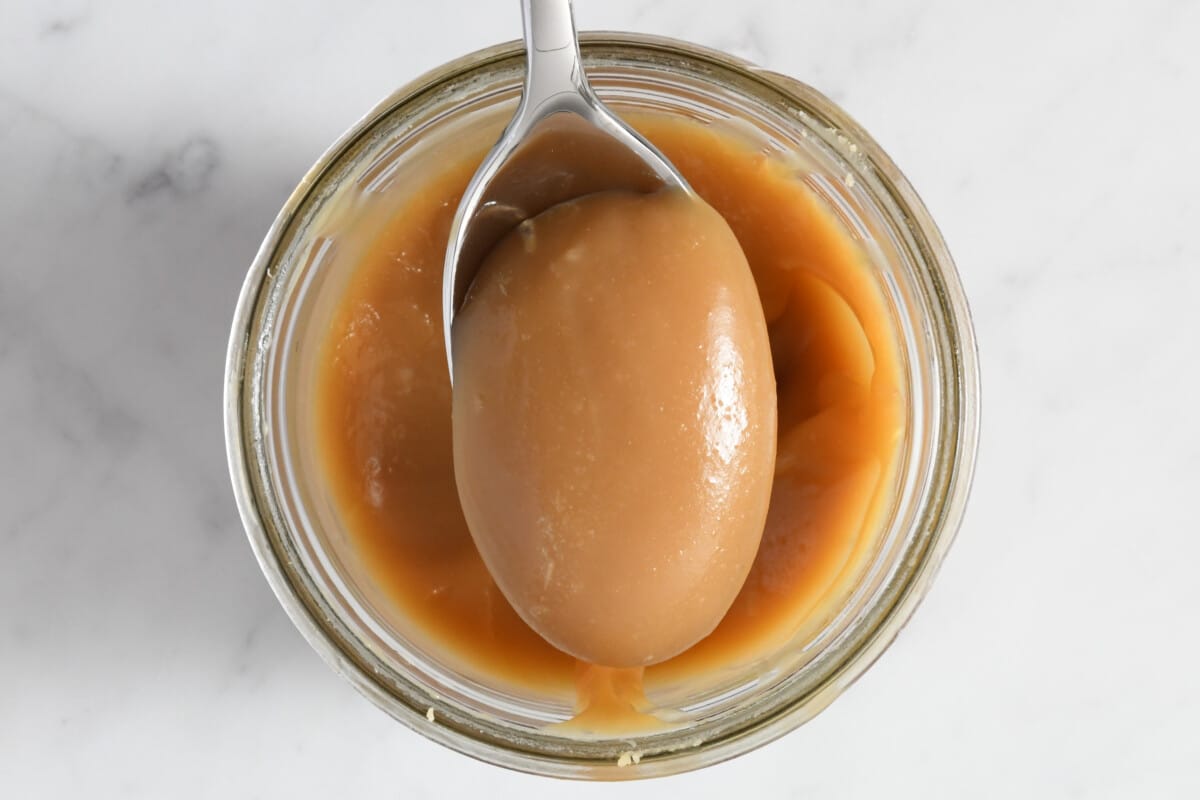 Metal spoon with a spoonful of caramel sauce in a glass jar on a marble counter.