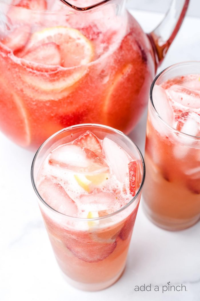 Glasses and a pitcher of strawberry lemonade with ice cubes and fresh strawberry and lemon slices.  // addapinch.com